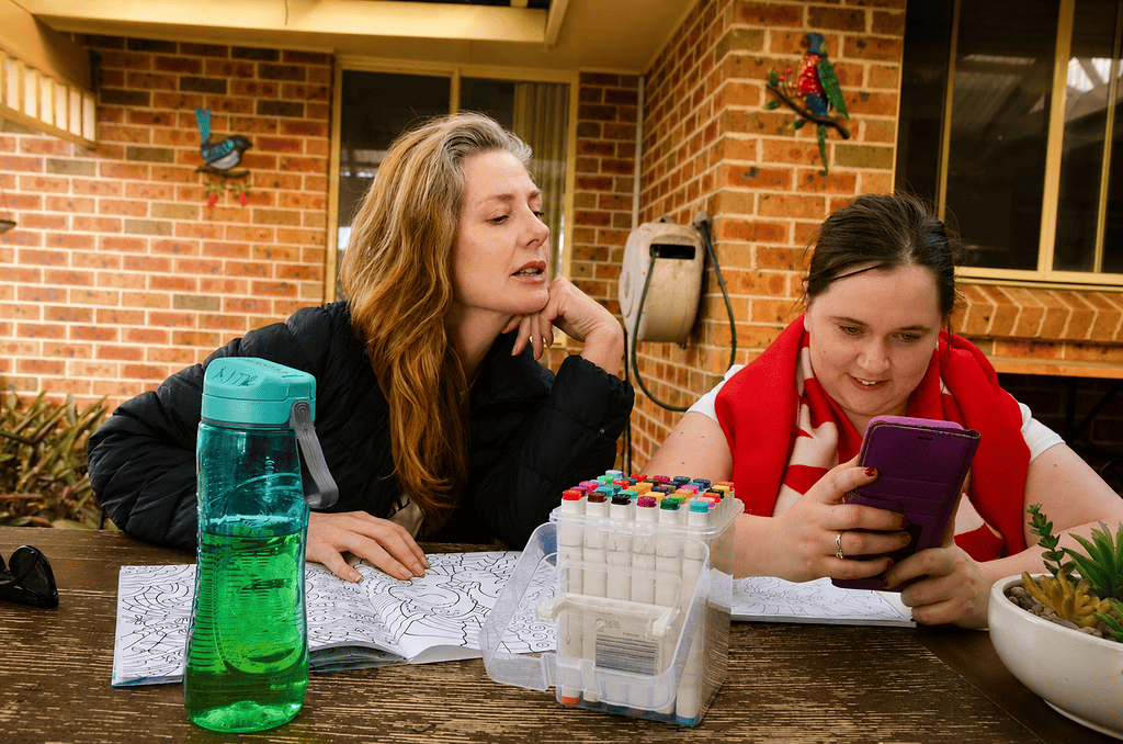 Two women are sitting at an outdoor table. One is looking at a mobile phone. The other is sneaking a peek over her shoulder.